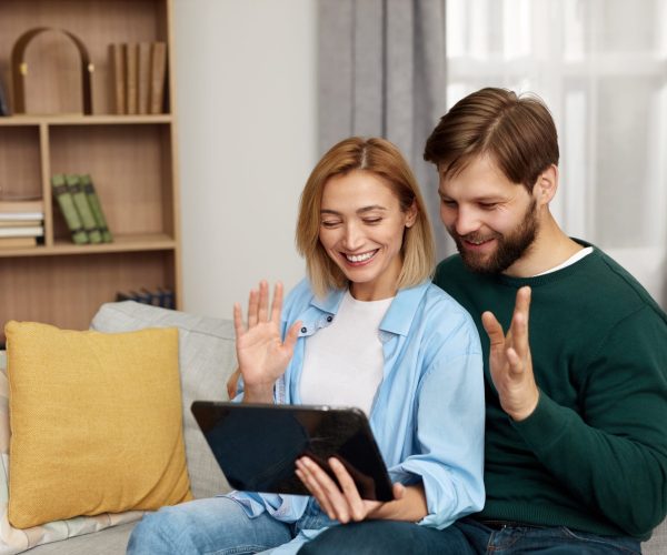 Couple Talking Tablet At Home. Young Couple Using Digital Tablet While Sitting On Couch. Happy Smiling People Watching Video On Digital Tablet At Home